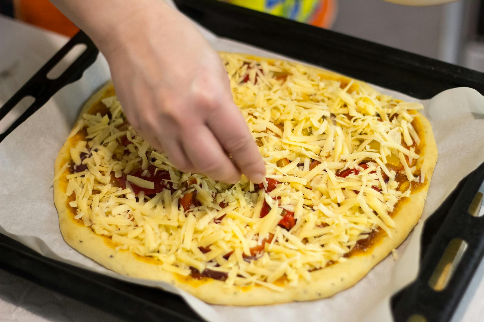Dłoń posypuje startym serem pizzę z sosem i dodatkami